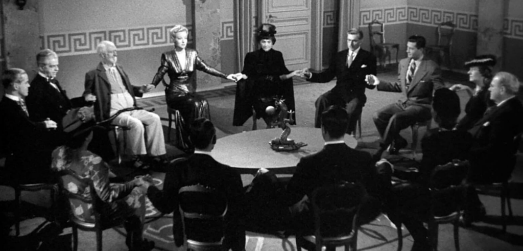 a circle of white people sitting around a round table with a dragon statue on it. The people are all dressed well and are holding hands.